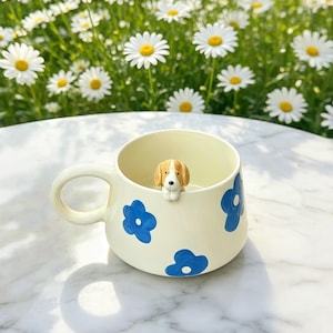 Op de afbeelding: Crèmekleurige keramische mok met blauwe bloemmotieven en een klein beagle-beeldje aan de binnenkant. De mok heeft een afgeronde vorm en een groot oor. Op de achtergrond zijn madeliefjes te zien.