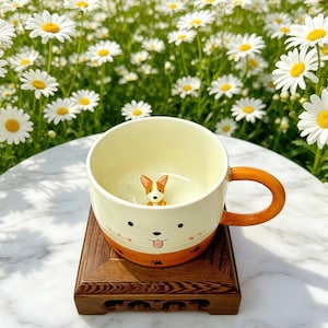Puede incluir: Una taza de cerámica color crema con un diseño de perro. Tiene un asa y una base de color naranja, con una cara de perro pintada en la parte delantera. Hay una pequeña figura de perro en el interior. La taza está sobre un soporte de madera.