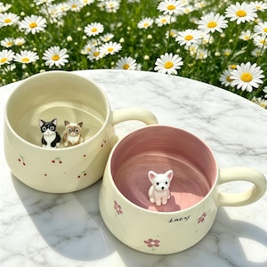 Puede incluir: Dos tazas de cerámica color crema con asas, decoradas con pequeñas flores rosas y cerezas. Una taza presenta una figura de gato blanco y negro y una figura de gato marrón. La otra taza tiene una figura de cachorro blanco y el nombre "Lacy".