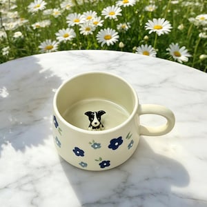 Könnte beinhalten: Ein cremefarbener Keramikbecher mit einem Muster aus kleinen dunkelblauen und hellblauen Blumen auf der Außenseite. Im Inneren des Bechers ist eine kleine, dreidimensionale schwarz-weiße Hundefigur am Boden befestigt, die nach oben blickt.