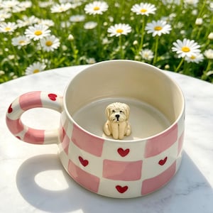 Könnte beinhalten: Ein Keramikbecher mit einem rosa-weißen Karomuster und kleinen roten Herzen. Der Henkel ist rosa-weiß gestreift und mit roten Herzen verziert. Im Inneren des Bechers befindet sich am Boden eine kleine, dreidimensionale cremefarbene Hundefigur.