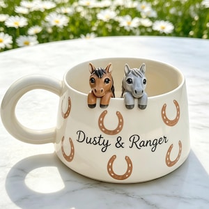 Puede incluir: Una taza de cerámica color crema con un patrón de iconos de herraduras marrones y el texto "Dusty & Ranger" en letra cursiva negra. Dos pequeñas figuras de caballos esculpidas, una marrón y otra gris, están fijadas al borde interior.