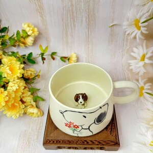 May include: Cream ceramic mug with a cartoon dog design in black and white, holding flowers. A small dog figurine is inside the mug. The mug has a handle and sits on a wooden stand, with yellow flowers in the background.