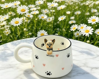 May include: A white ceramic mug with a Yorkshire Terrier figurine perched on the rim. The mug features black paw prints and pink hearts, with the name "Whiskey" printed on the side. The background is a field of white daisies.
