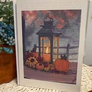 May include: Greeting card featuring a watercolor painting of a lantern with a lit candle, pumpkins, and fall foliage. The lantern is black with a glass enclosure. The pumpkins are orange, and the foliage includes sunflowers and other autumn flowers.