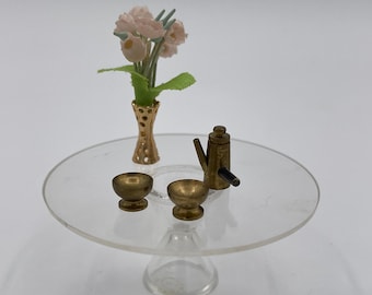 May include: Miniature dollhouse table setting with a clear glass table, gold-colored vase with pink flowers, two small gold bowls, and a gold-colored teapot. The table is round and has a clear pedestal base.