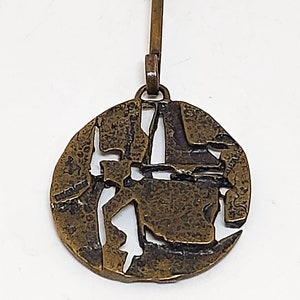 May include: A bronze pendant necklace with a circular design featuring a textured, abstract pattern with cut-out sections. The pendant is attached to a chain with a simple clasp.