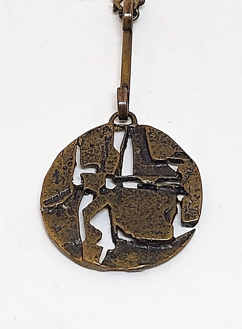 May include: A bronze pendant necklace with a circular design featuring a textured, abstract pattern with cut-out sections. The pendant is attached to a chain with a simple clasp.
