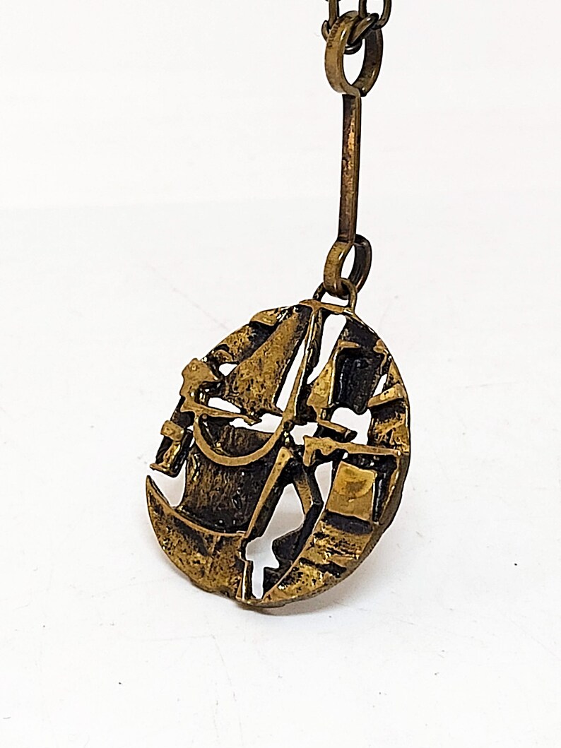 May include: A gold-toned pendant necklace featuring a stylized abstract design. The pendant is shaped like a circle with a textured surface and intricate details.