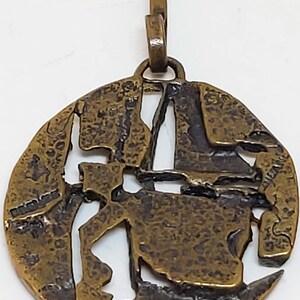 May include: A round, bronze pendant with a textured surface and a cutout design. The pendant has a loop at the top for attaching to a chain.