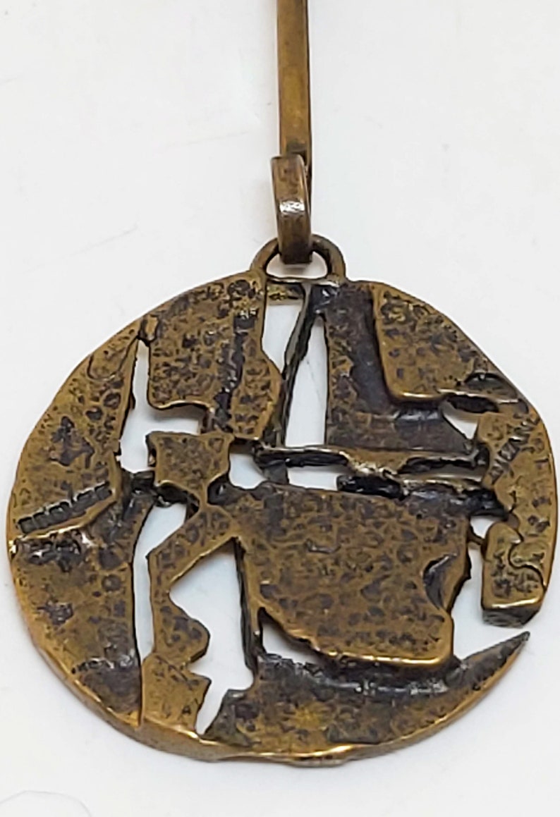 May include: A round, bronze pendant with a textured surface and a cutout design. The pendant has a loop at the top for attaching to a chain.