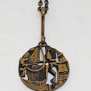 May include: A gold-toned necklace with a round pendant featuring an abstract design. The pendant is made of metal with a textured surface and has a cutout design.