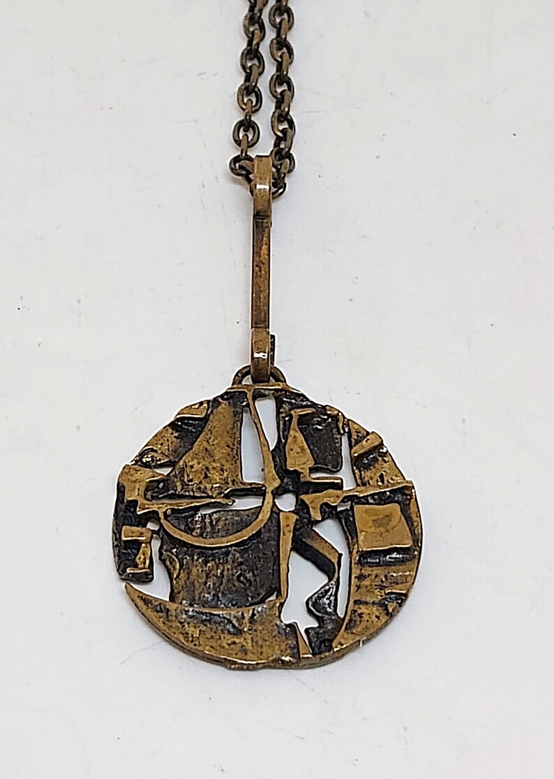May include: A gold-toned necklace with a round pendant featuring an abstract design. The pendant is made of metal with a textured surface and has a cutout design.