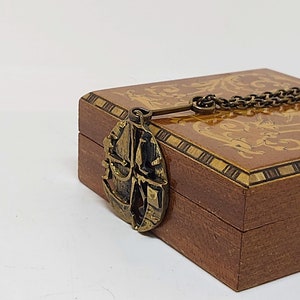 May include: A wooden box with a decorative inlay design and a brass pendant attached to a chain. The pendant features a stylized ship design.