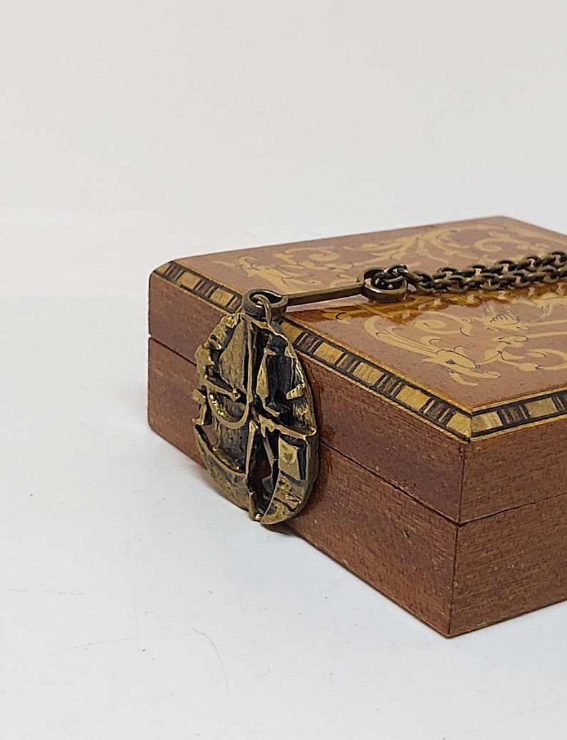 May include: A wooden box with a decorative inlay design and a brass pendant attached to a chain. The pendant features a stylized ship design.