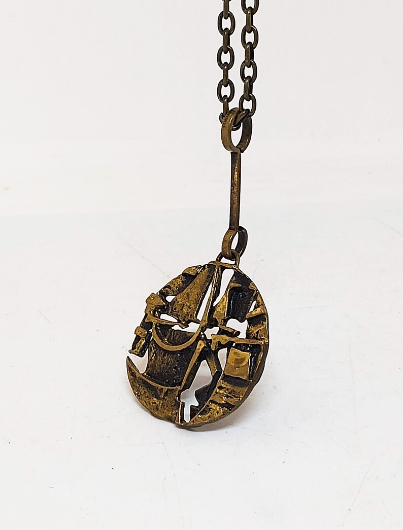 May include: A gold-toned necklace with a pendant featuring a detailed design of a sailboat with a rope and bucket. The pendant is round and has a textured surface.