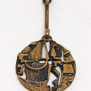 May include: A gold-toned pendant necklace with a circular, abstract design. The pendant features a textured, geometric pattern with cutouts.