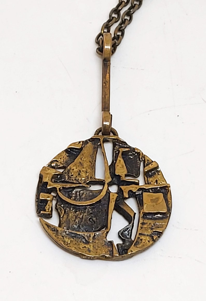 May include: A gold-toned pendant necklace with a circular, abstract design. The pendant features a textured, geometric pattern with cutouts.