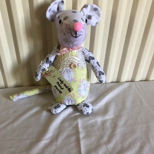 May include: A handmade plush toy mouse with a pink nose and grey polka dots on its ears. The mouse is wearing a light green and white floral dress with a pink bow. The mouse is holding a yellow tag that says "Lady and the Tramp Mouse".