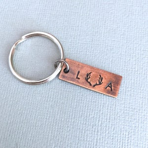 May include: A copper keychain with a silver key ring. The keychain has the letters "L" and "A" on either side of a pair of antlers.