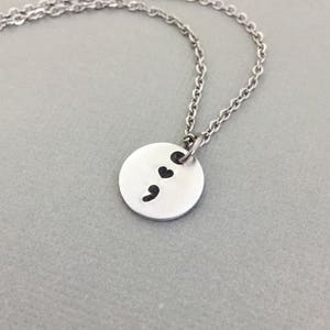 Pode incluir: Um colar de prata com um pingente redondo que apresenta um ponto e vírgula e dois pequenos corações. O colar está sobre um fundo branco.