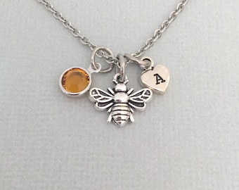 May include: A silver necklace with a bee charm, a small amber-colored gemstone, and a heart charm with the letter "A" engraved on it.