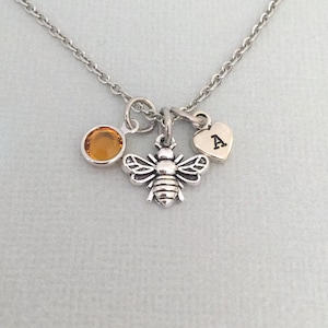 May include: A silver necklace with a bee charm, a small amber-colored gemstone, and a heart charm with the letter "A" engraved on it.