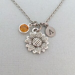 May include: Silver necklace with a sunflower charm, a small orange crystal, and a silver charm with the letter A.