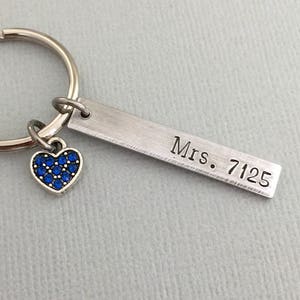 Könnte beinhalten: Ein silberner Schlüsselanhänger mit einem kleinen blauen Strass-Herz-Anhänger und einem rechteckigen Anhänger, der mit "Mrs. 7125" graviert ist.