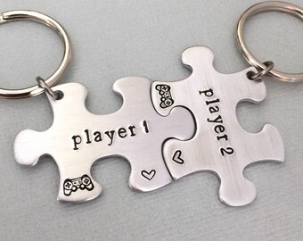 Op de afbeelding: Twee zilveren puzzelstukjes sleutelhangers met de woorden "Player 1" en "Player 2" erop gegraveerd. Elk stuk heeft een klein hartje en een symbool van een videogamecontroller.