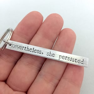 Könnte beinhalten: Silberner Schlüsselanhänger mit einem rechteckigen Anhänger, auf dem "nevertheless, she persisted" steht.