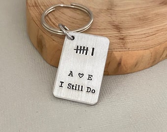 Op de afbeelding: Zilveren sleutelhanger met een rechthoekig label waarop "A ♥ E I Still Do" staat en een rij van vijf hash-markeringen boven de tekst.