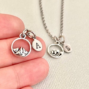 May include: Two silver necklaces with charms. One charm features a mountain range inside a circle, the other charm features a wave inside a circle. Both charms have a small circle with a letter on them. The letter on the mountain charm is "A" and the letter on the wave charm is "S".