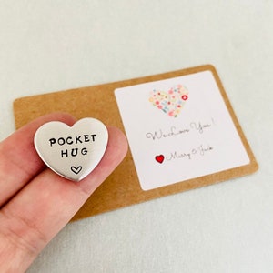 May include: A silver heart-shaped token with the words "Pocket Hug" and a small heart symbol. The token is being held in a hand and is accompanied by a card with a floral heart design and the text "We love you!" with the names "Marry & Jack" underneath.