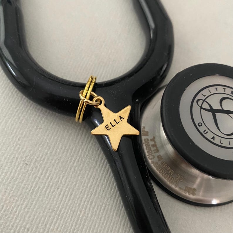 May include: Gold star-shaped charm with the name "ELLA" engraved on it. The charm is attached to a stethoscope with a black tube and a silver-colored head.