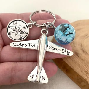 May include: Silver airplane keychain with a compass charm and a glass globe charm. The airplane has the words "Under The Same Sky" engraved on it.