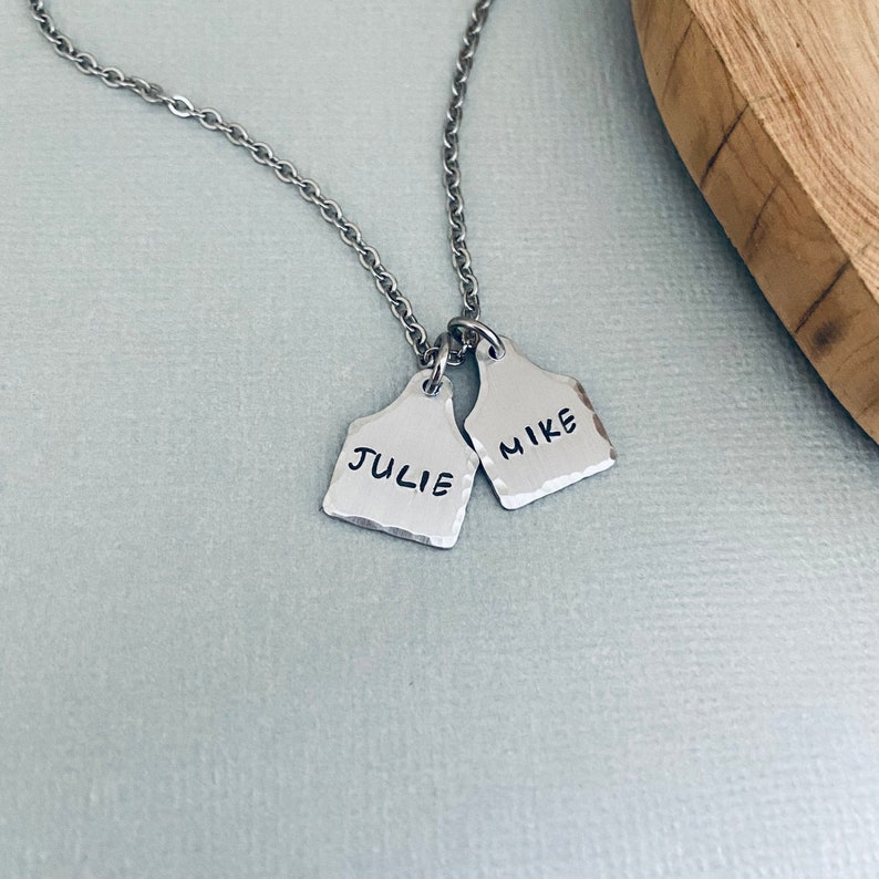 Op de afbeelding: Een zilveren ketting met twee zilveren hangers. De hangers zijn gevormd als een koe-hanger en hebben de namen "Julie" en "Mike" erop gegraveerd.