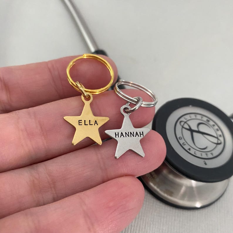 May include: Two gold and silver star-shaped keychains with the names "Ella" and "Hannah" engraved on them. The keychains are attached to a stethoscope.