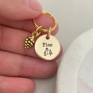 May include: Gold keychain with a pine cone charm and a round tag that says "Pine" with a small image of three pine trees.