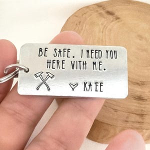 以下が含まれることがあります： 「BE SAFE. I NEED YOU HERE WITH ME.」のテキストとハートマークの下に「KATE」という名前が入った長方形のシルバーキーホルダー。 キーホルダーには交差した斧も付いています。