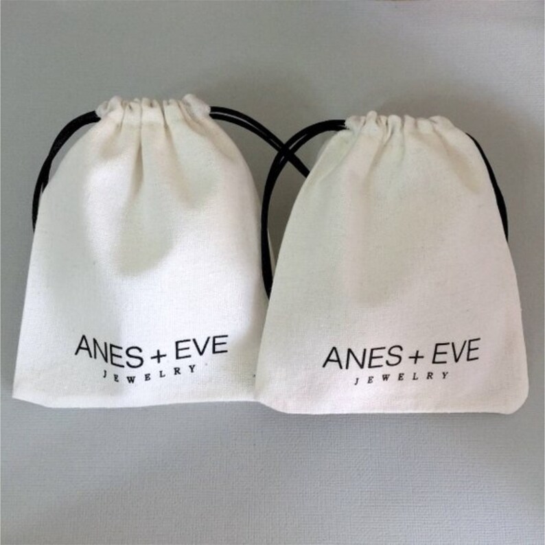Peut inclure: Deux sacs en coton blanc avec des cordons de serrage noirs. Les sacs portent l'inscription "ANES + EVE JEWELRY" imprim&eacute;e sur le devant.
