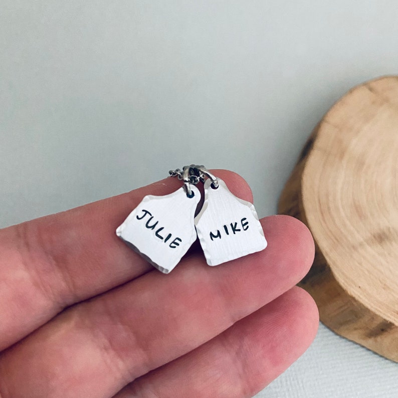 Op de afbeelding: Zilveren ketting met twee gepersonaliseerde tags. De tags zijn gevormd als oorkleppen voor vee en hebben de namen "Julie" en "Mike" erop gegraveerd.