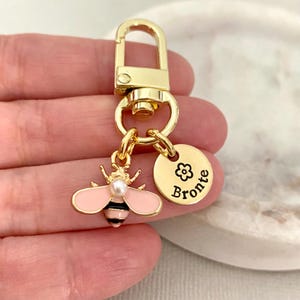 Puede incluir: Llavero dorado con un dije de abeja rosa y negra y una etiqueta redonda dorada con el nombre "Bronte" grabado.