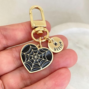 Puede incluir: Llavero dorado con un dije en forma de corazón negro con un diseño de telaraña y un dije redondo con la palabra "Willow" y un pequeño gráfico de araña.
