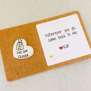 Peut inclure: Un petit jeton en forme de c&oelig;ur argent&eacute; avec les mots "One Day Closer" et une petite &eacute;tiquette avec les lettres "AOE" dessus. Une carte blanche avec le texte "Wherever you go... come back to me" et un c&oelig;ur rouge avec le nom "Kat" &eacute;crit en dessous.