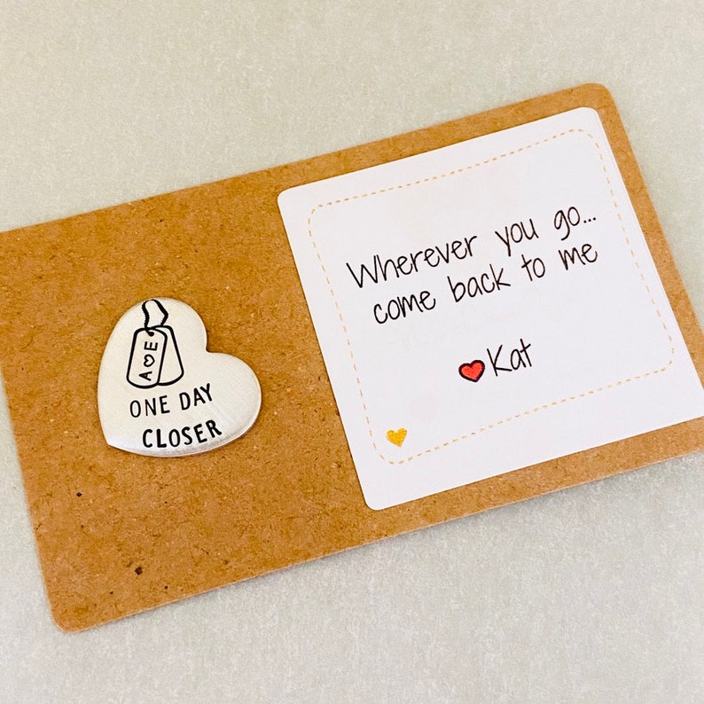 Peut inclure: Un petit jeton en forme de c&oelig;ur argent&eacute; avec les mots "One Day Closer" et une petite &eacute;tiquette avec les lettres "AOE" dessus. Une carte blanche avec le texte "Wherever you go... come back to me" et un c&oelig;ur rouge avec le nom "Kat" &eacute;crit en dessous.