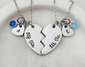 May include: Two silver necklaces with heart pendants that say "Big Sis" and "Lil Sis". Each necklace has a small heart charm with a letter and a blue gemstone.