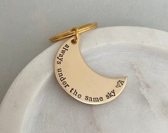 May include: A gold-colored crescent moon keychain with the words "always under the same sky" engraved on it. The keychain has a small ring attached for keys. The background is a white marble surface.