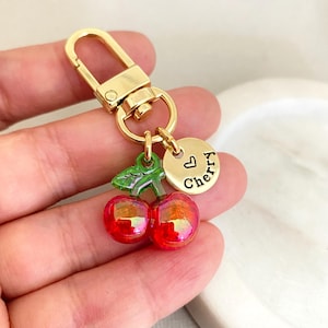 Puede incluir: Llavero dorado con un dije de cereza roja y verde y un dije dorado con la palabra "Cherry" y un símbolo de corazón.
