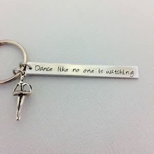 Op de afbeelding: Zilveren sleutelhanger met een klein danser-bedeltje en een zilveren staaf met de inscriptie "Dance like no one is watching."